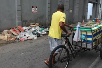 Homem conduz triciclo de carga por uma rua, ao lado de resíduos descartados junto a um muro.